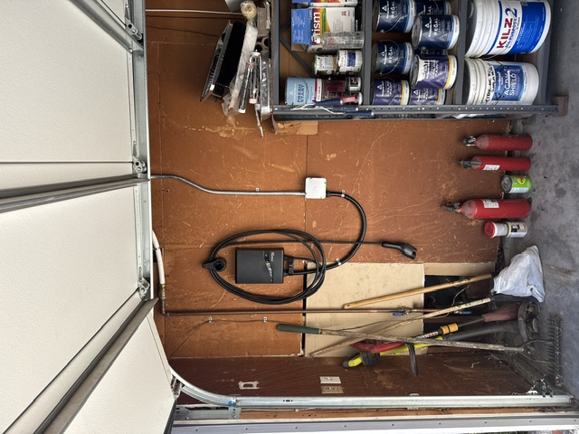 A garage wall with a mounted EV charger and conduit run above a shelf of paint cans