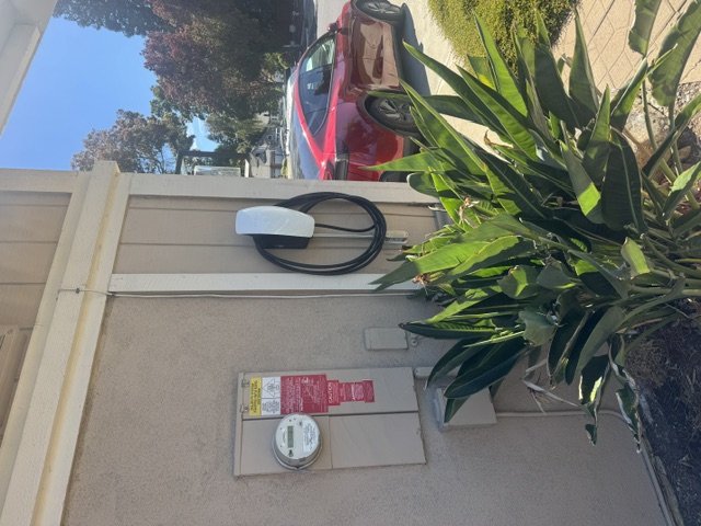 An EV charger mounted on a stucco exterior wall beside a meter, with a red car parked in front