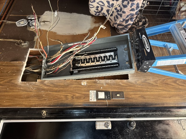 An old panel being replaced, with loose wiring exposed on a wooden wall beside a blue ladder
