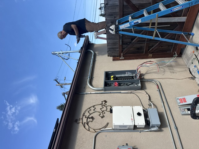 An electrician on a ladder working at an exterior panel beside a solar inverter under a blue sky