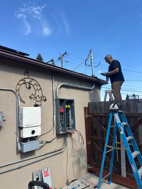 An electrician on a blue ladder wiring an exterior electrical panel beside a solar inverter on a stucco wall